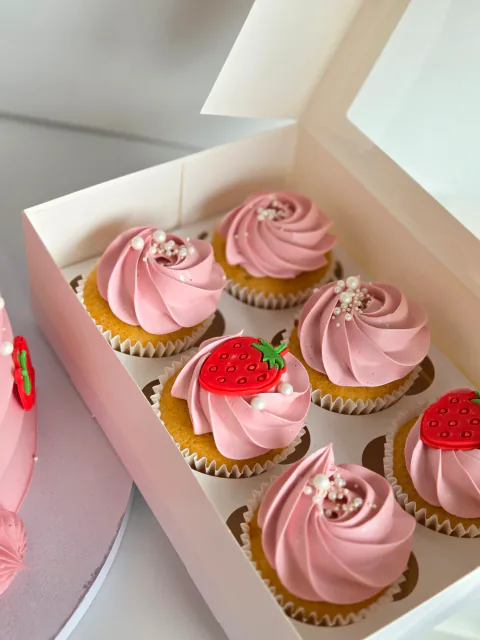 Pink strawberry cupcakes with buttercream swirls beside a matching celebration cake